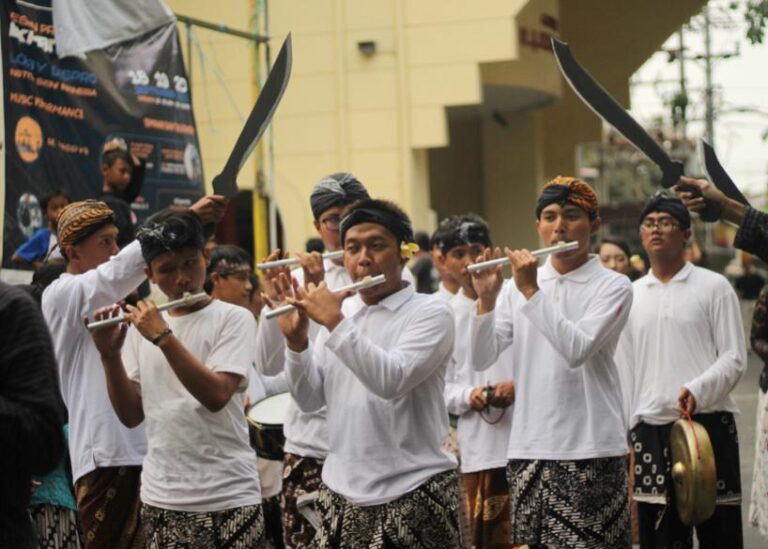 Group of young men in white shirts and batik skirts playing flutes in a street parade with onlookers and banners behind them.